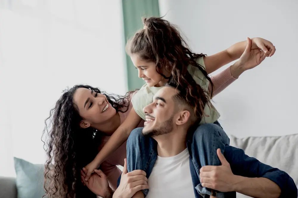 two adults smiling with a child seated on one adult’s shoulders