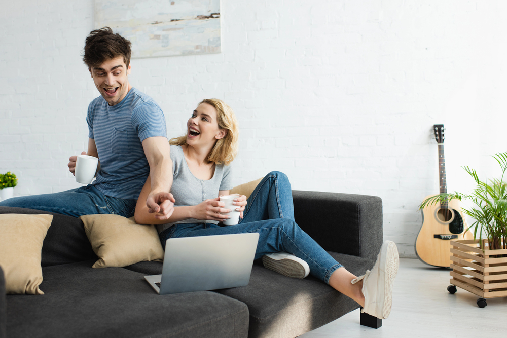 What to Expect at a Luxury Addiction Treatment Facility in California 7 Happy couple on sofa with laptop, guitar, and plant—reflecting a calm, home-like luxury addiction treatment center.
