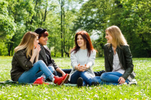 Four friends sit on sunny grass in a park, laughing and chatting together outdoors.