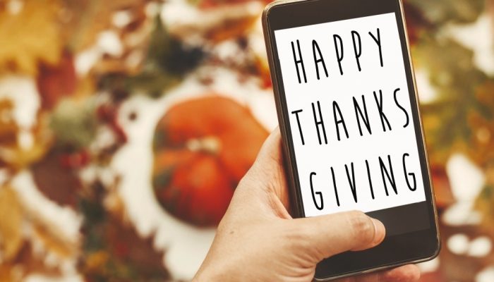 happy thanksgiving text on phone screen in hand at stylish autumn flat lay of pumpkin in leaves wreath.